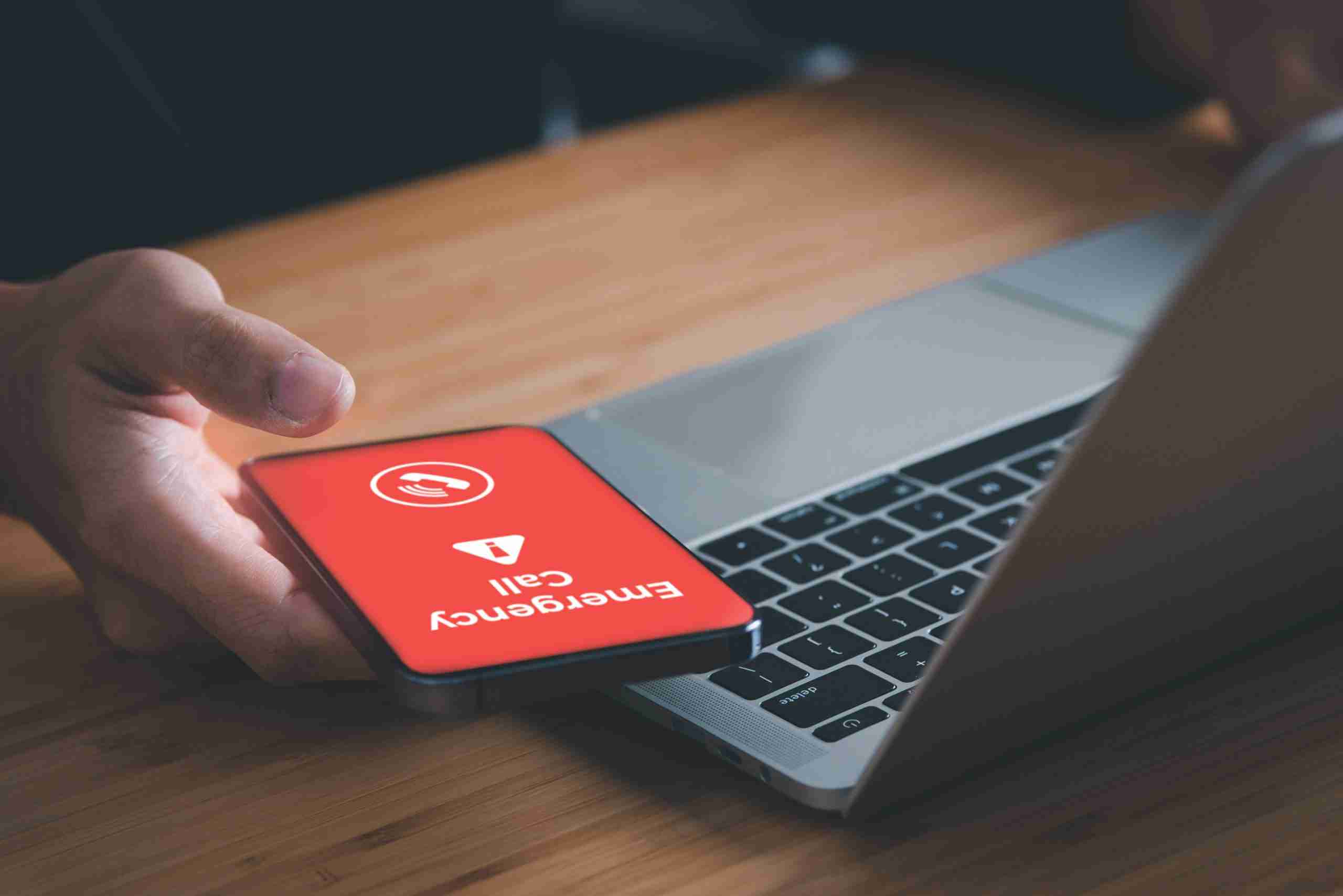 man using a laptop and holding a phone with emergency message on it